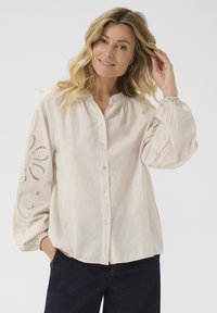 Chemise boutonnie beige clair avec col mandarin, présentant de longues manches bouffantes et des détails en dentelle élaborés sur la manche gauche.