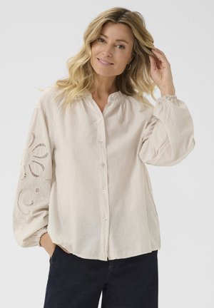 Chemise boutonnie beige clair avec col mandarin, présentant de longues manches bouffantes et des détails en dentelle élaborés sur la manche gauche.