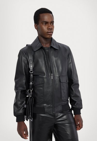 Čierna kožená bomber bunda so zipsom vpredu, klasickým golierom, dvoma prednými vreckami a rebrovanými manžetami. Model nesie zodpovedajúcu tašku.
