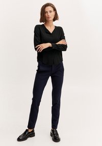 Blusa nera a maniche lunghe con scollatura a V e dettagli con bottoni, abbinata a pantaloni slim-fit blu navy e mocassini neri. Design semplice e pulito.