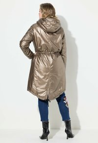Chaqueta larga de metal bronce con cintura ajustable, capucha y dobladillo curvado, combinada con unos vaqueros azules oscuros con bordado floral.