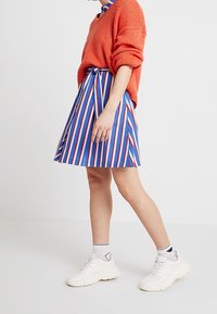 Gestreifter Rock in Blau, Rot und Weiß mit A-Linien-Design, kombiniert mit einem orangen gestrickten Pullover und weißen Sneakers.