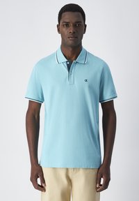 Jasnoniebieska koszulka polo z bawełnianego materiału, z granatowym kołnierzykiem, białymi obszyciami oraz małym logo na piersi. Krótkie rękawy.