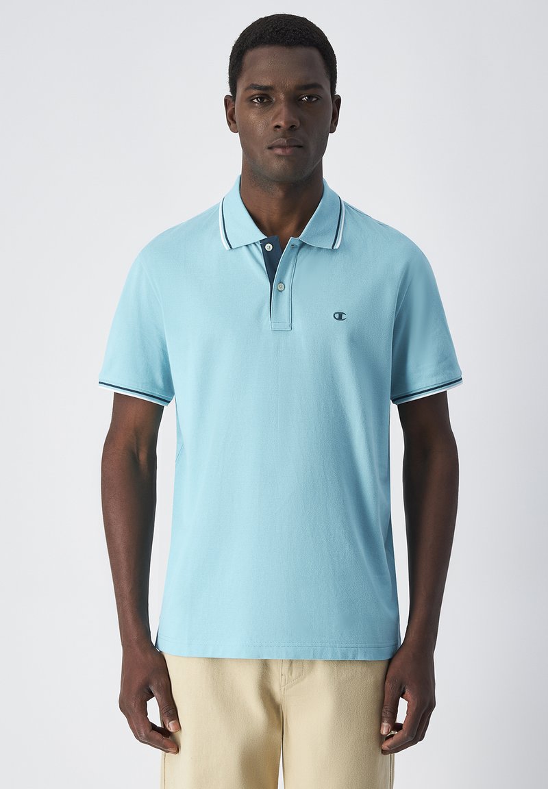 Jasnoniebieska koszulka polo z bawełnianego materiału, z granatowym kołnierzykiem, białymi obszyciami oraz małym logo na piersi. Krótkie rękawy.