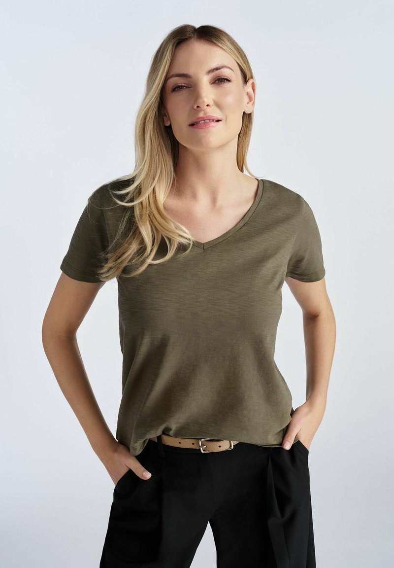 Oliwkowa koszulka z dekoltem w serek w miękkim, teksturowanym materiale, z krótkimi rękawami, zestawiona z czarnymi spodniami i jasnobrązowym paskiem.