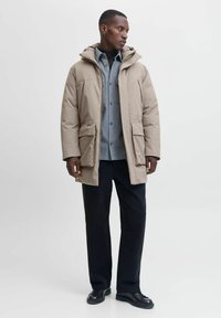 Bežna izolirana parka s kapuco, dvema sprednjima žepoma in zadrgo, nošena čez sivo srajco, dopolnjena z temnimi hlačami in čevlji.