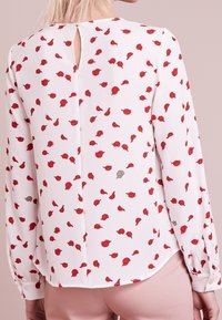 Femme portant un chemisier blanc à manches longues avec un motif floral rouge et une ouverture en clé à l'arrière, associé à un pantalon rose clair.