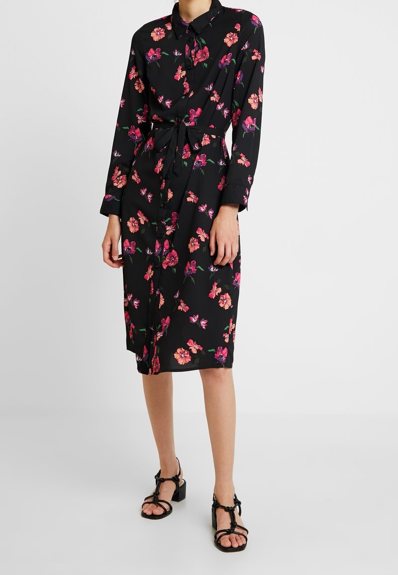 Robe noire avec des motifs floraux roses et violets, manches longues, col, boutons à l'avant, et une ceinture assortie. Portée avec des sandales noires à talons.