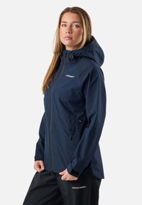 Žena oblečená v tmavomodrej nepremokavej bunde s kapucňou a čiernych nohaviciach, stojaca s rukami pri vreckách, neutrálneho výrazu, na jednoliatom pozadí.