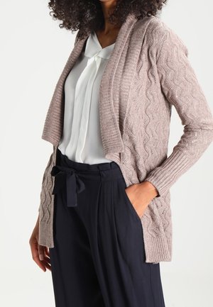 Femme portant un cardigan tricoté marron clair sur un chemisier blanc rentré dans un pantalon noir taille haute avec une ceinture en tissu nouée.