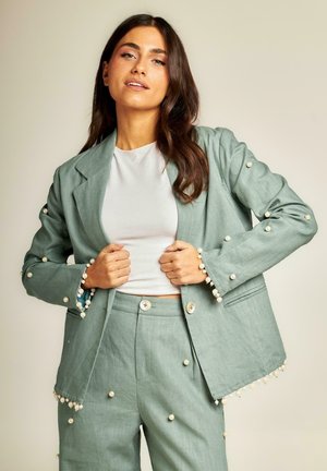 Traje de lino verde claro con acentos de perlas en las mangas y el dobladillo. Cuenta con un blazer entallado y pantalones a juego con cierre de botón.