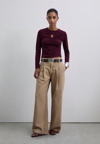 Burgundy long-sleeve top with a keyhole detail, paired with beige wide-leg trousers and brown sandals adorned with studs. Silver cuff bracelet visible.