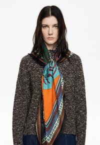 Bufanda de seda con acentos en turquesa, naranja y un diseño estampado, drapeada elegantemente alrededor del cuello, combinada con un suéter marrón texturizado.