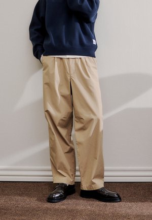 Persona che indossa pantaloni beige a gamba larga, mocassini neri in pelle e una felpa blu navy, in piedi contro un muro chiaro con ombra.