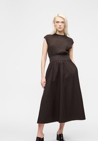 Γυναίκα με ανοιχτά μαλλιά που φοράει αμάνικο midi φόρεμα σε σκούρο καφέ με φαρδιά ζώνη στη μέση και μαύρα παπούτσια με τακούνι χωρίς λουριά, στέκεται μπροστά σε λευκό φόντο.