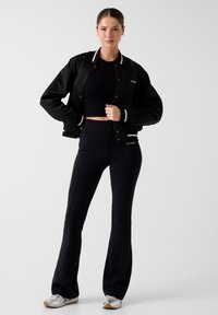 Giacca bomber nera con accenti bianchi, top corto aderente nero e pantaloni neri svasati, indossati con sneakers argento con il logo del marchio.