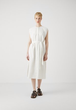 Robe blanche mi-longue en tissu texturé, à manches courtes, avec une fermeture à boutons sur le devant et une ceinture en tissu assortie. Portée avec des sandales à lanières marron foncé.