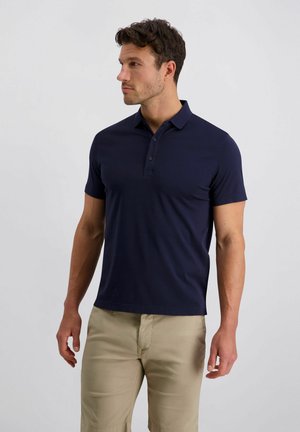 Homme debout portant un polo bleu marine à manches courtes et un pantalon beige, regardant vers sa gauche sur un fond uni.