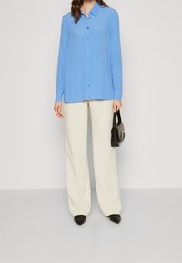 Camicia a maniche lunghe blu chiaro con chiusura a bottone, abbinata a pantaloni beige a gamba larga e tacchi neri a punta. Inclusa una piccola borsa nera.