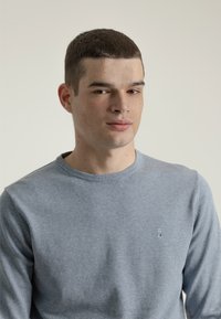 Suéter de cuello redondo azul claro con mangas largas, textura suave y pequeño logo bordado en el pecho. Diseño liso y ajustado.