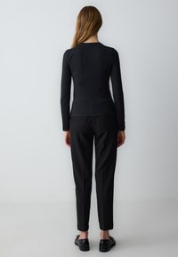 Schwarzes Langarmoberteil mit taillierter Silhouette, kombiniert mit schwarzen schmal zulaufenden Hosen und schwarzen Slippers, von hinten betrachtet.