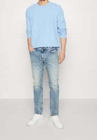 Ljust blå crew-neck tröja med åtsittande ärmar, kombinerad med bleka blå jeans. Vita sneakers med texturerad detalj. Enkel, ren design.