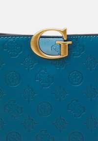 Superficie in pelle blu impresso con motivi geometrici ripetuti e una grande lettera "G" metallica dorata attaccata al bordo superiore.
