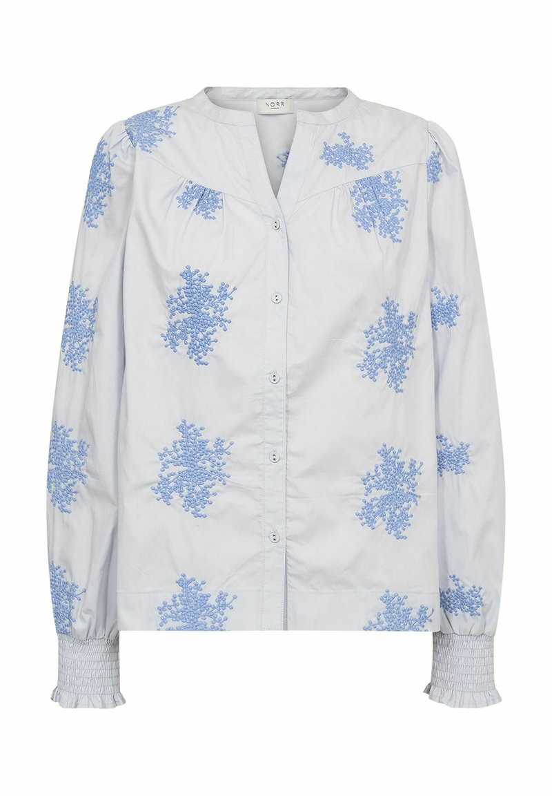 Camicia azzurra chiaro con bottoni in tessuto bianco, caratterizzata da ricami floreali blu a rilievo e polsini arricciati. Design con colletto classico.