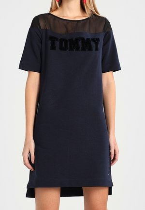 Kobieta ubrana w granatową sukienkę z krótkim rękawem, z przezroczystym czarnym panelem na górze i napisem „TOMMY” na piersi, zestawiona z czerwonymi kolczykami.