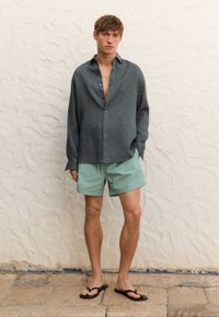 Camicia grigia in lino a maniche lunghe sbottonata al colletto, abbinata a pantaloni corti verde chiaro e infradito nere. Sfondo di una parete bianca texturizzata.
