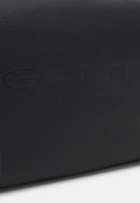 Musta teksturoitu nahkalaukku, jossa on kohokuvioitu brändimerkiksi, esillä "GANT" ja "EST. 1949" -yksityiskohdat.