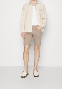 Camisa beige de botones sobre una camiseta blanca sin mangas, combinada con pantalones cortos marrón claro y sandalias crema con correa, sobre un fondo neutro.