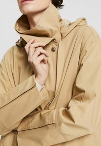 Persona che indossa una giacca beige con colletto alto, mentre tiene il colletto con una mano vicino al viso, tagliata sopra le labbra.