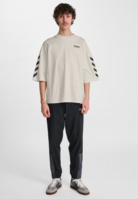Crème kleur t-shirt met korte mouwen, voorzien van een zwart logo en strepen op de mouwen, gecombineerd met zwarte joggers met een klein logo en grijze paneelaccenten.