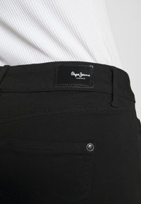 Jeans de mezclilla negra con una etiqueta de cuero en la cintura que presenta "Pepe Jeans London." Incluye un acento metálico en el bolsillo y detalles de costura sutiles.