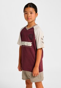 Vinröd sportströja med ljusbeige ärmar och design med V-ringning. Har en logotyp och texten "BEES" i vitt. Matchas med beige shorts.