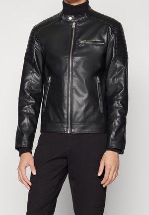 Czarna kurtka motocyklowa z czarnej sztucznej skóry z przednim zamkiem błyskawicznym, zapinanymi na zamek kieszeniami na piersi i po bokach, noszona na czarnym golfie i czarnych spodniach.