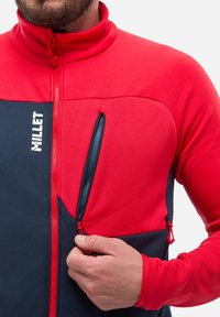 Rote und marineblaue Fleecejacke mit Reißverschlusstaschen, hohem Kragen und kontrastierenden Texturen; mit einem Logo auf der linken Brust.