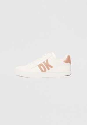 Fehér sneaker sima textúrával, oldalt egy figyelemfelkeltő világos rózsaszín "DK" logóval és a saroknál egy tompított rózsaszín kiegészítéssel.