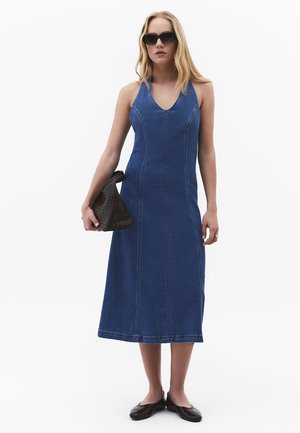 Donna che indossa un vestito midi di denim blu senza maniche, scarpe nere basse, occhiali da sole neri e tiene una pochette nera intrecciata.