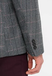 Manica di un blazer grigio a scacchi con un tessuto testurizzato. I dettagli più significativi includono tre bottoni neri e sottili accenti rosa nel motivo.