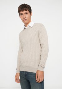 Beige ribbestrikket genser med rund hals, lange ermer og teksturert overflate, båret over en hvit skjorte med krage og kombinert med blå jeans.