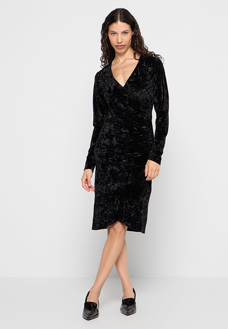 Schwarzes Samt-Wickelkleid mit langen Ärmeln, V-Ausschnitt und knielanger Hemdlänge. Es hat eine dezente Textur und eine taillierte Silhouette. Abgerundet mit glänzenden schwarzen Schuhen.