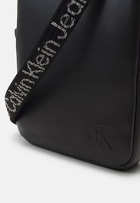 Borsa a tracolla in pelle nera con superficie texturizzata, dotata di una tracolla intrecciata con la scritta "Calvin Klein Jeans" e un logo "CK" in rilievo.