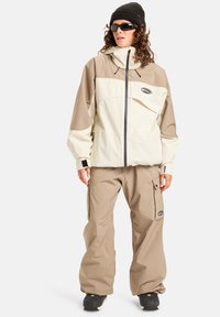 Beige og krem vanntett jakke med justerbar hette, glidelås, og logo. Kombinert med matchende beige cargo-bukser. Svart lue og solbriller.
