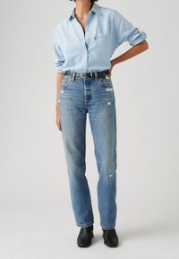 Ljusblå denimskjorta med en bröstficka, kombinerad med slitna blå jeans med raka ben, slitna områden och ett svart bälte.