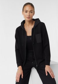 Schwarzer Reißverschluss-Sweatshirt mit Kapuze, ausgestattet mit einer Fronttasche. Glatter Stoff und lässige Passform. Getragen über einem schwarzen Tanktop und einer Hose.