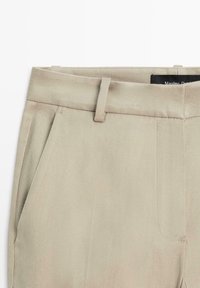 Světlé béžové kalhoty s vysokým pasem, poutky na opasek, předními záhyby, bočními kapsami a viditelnou černou značkovou etiketou uvnitř pásu.