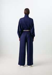 Casaco azul-marinho, curtido, com gola alta e mangas bufantes, combinado com calças de perna larga com cintura elástica. Textura suave.