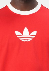 Κόκκινο βαμβακερό T-shirt με λευκό λαιμόκοψη. Διαθέτει προεξέχον λευκό λογότυπο τριφυλλιού Adidas στο στήθος με τρεις οριζόντιες ρίγες.